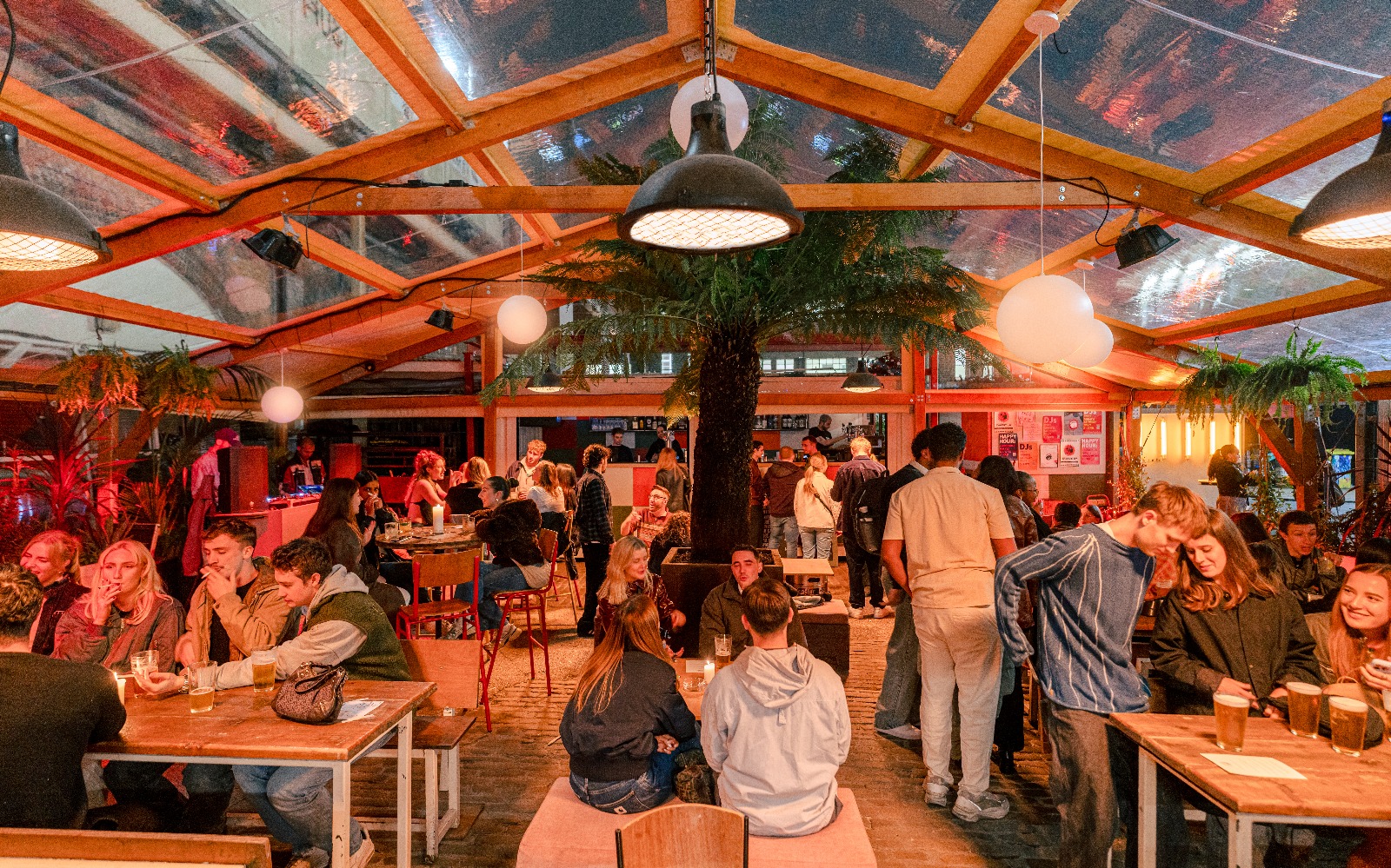 Peckham Arches covered terrace in the evening, decorated with globe lights, tropical ferns and plants. Guests socialise at tables beneath a clear roof.
