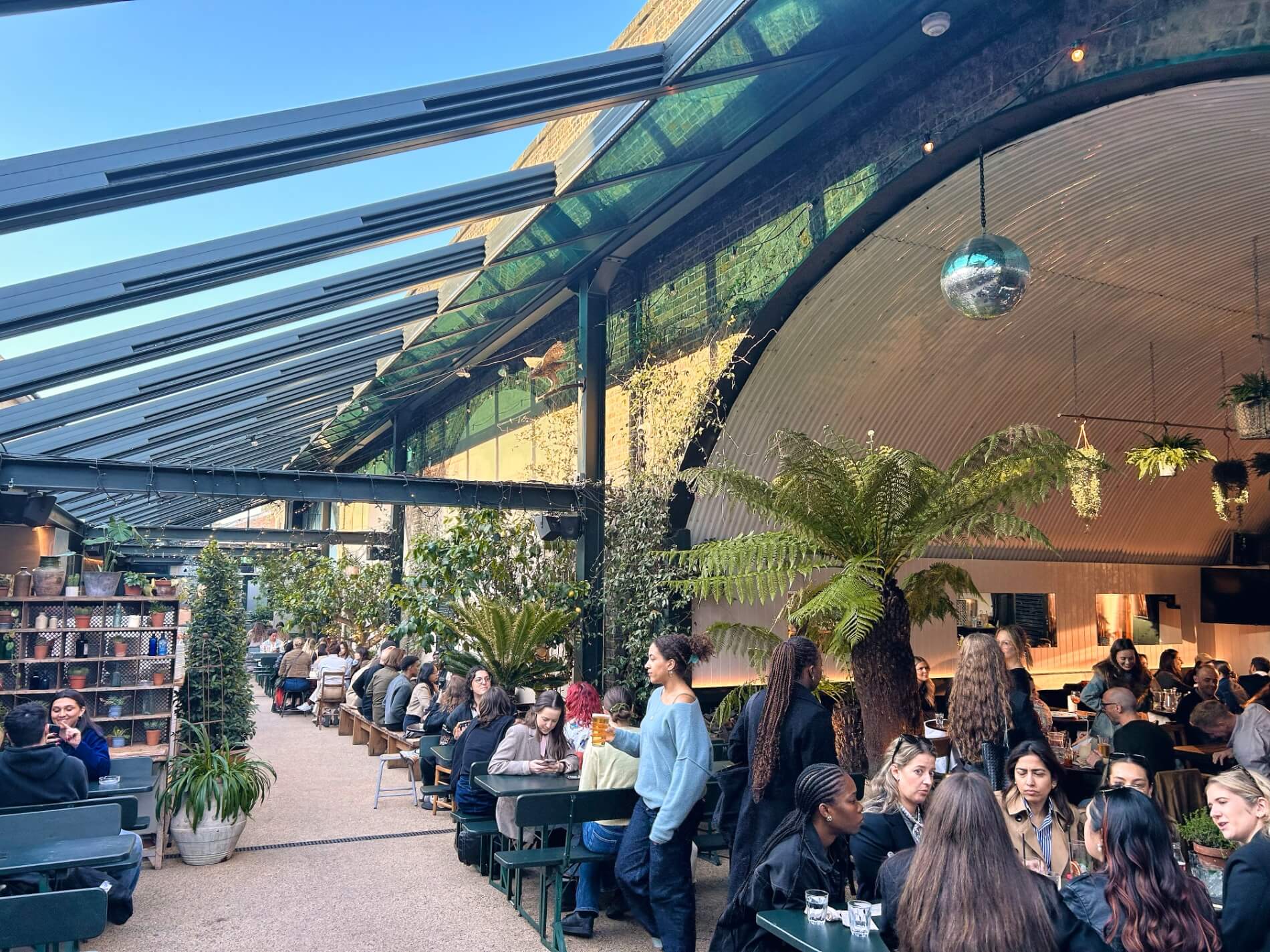 Guests at The Hawk's Nest dining in a warmly lit glass covered roof interior decorated with hanging greenery, fairy lights and flowers.