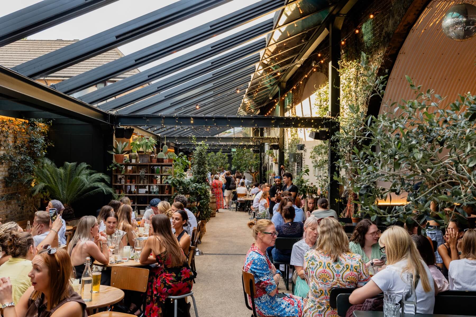 Guests at The Hawk's Nest dining in a warmly lit glass covered roof interior decorated with hanging greenery, fairy lights and flowers.